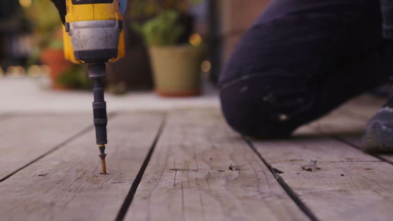 Close-up of a yellow drill driving a screw into a wooden plank, with a blurred background of potted plants and a person kneeling nearby.
