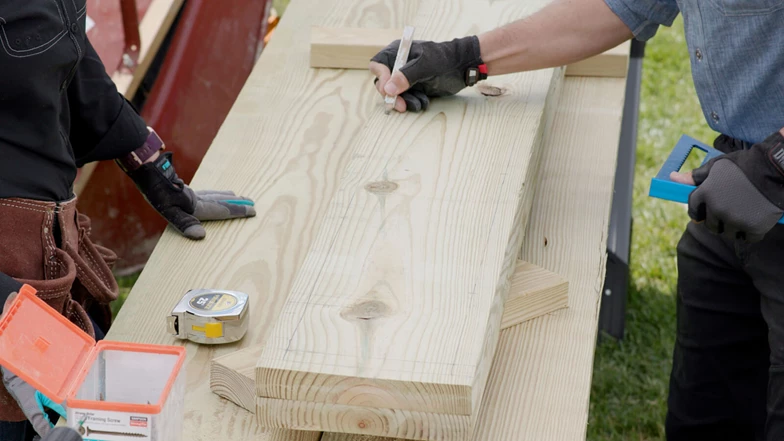 2 standing over a board. 1 person is marking the board with a carpenters pencil.